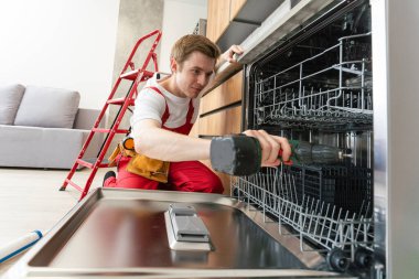 Üniformalı teknisyen ya da işçi mutfak mobilyalarına bulaşık makinesi yerleştiriyor. Tamirci bulaşık makinesini tamir ederken işçi kıyafeti giyer. Koruyucu eldivenlerde uzman Bulaşık makinesini tamir eder.