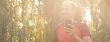 Tişörtlü yakışıklı bir adam parkta antrenman yapıyor ve kulaklıkla müzik dinliyor..