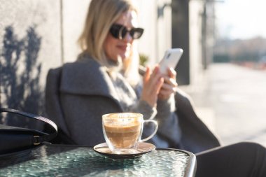 Ön planda bir fincan latte ile bronzlaşmış kadının portresi. Kahvaltının ortasında güneş gözlüklü gülümseyen kadın. Yüksek kalite fotoğraf