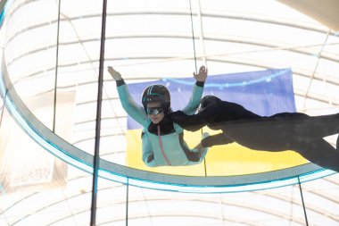 Excited teenage girl in helmet flying in aerodynamic tube wind tunnel. Skydiving training. High quality photo clipart