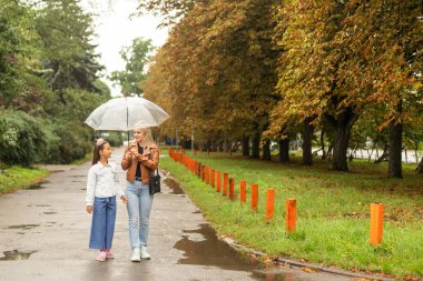 Cheerful mother and her little daughter having fun together in the autumn background under the umbrella. Happy family in the fall background.