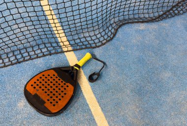 Bir padel veya raket tenis raketi ve parlak sarı bir tenis topu mavi bir kortta yatar. Ağ ve bir oyuncunun bulanık figürü arka planda görünür. Yüksek kalite fotoğraf