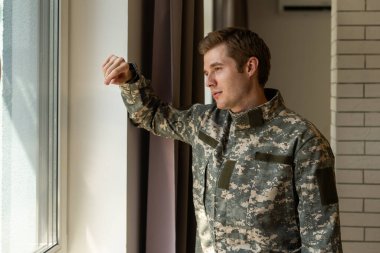 Serious short-haired young man in military uniform soldier looking through the window, upset soldier suffering from posttraumatic syndrome after returning home, copy space, closeup