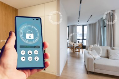 Elinde akıllı telefon tutan ve evinde güvenlik uygulaması olan genç bir kadın. Ev güvenliğini mobil bir cihazdan kontrol etme ve yönetme kavramı.