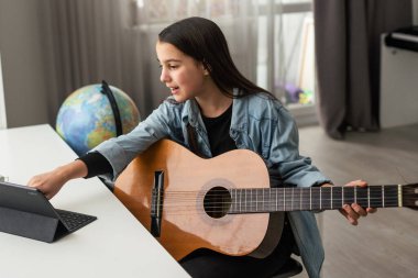 Genç kız online dersleri kullanarak evde gitar çalmayı öğreniyor. Uzaktan hobi müzik eğitimi akustik gitar. Uzayı kopyala Yüksek kalite fotoğraf
