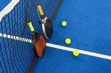 Bir padel veya raket tenis raketi ve parlak sarı bir tenis topu mavi bir kortta yatar. Ağ ve bir oyuncunun bulanık figürü arka planda görünür. Yüksek kalite fotoğraf