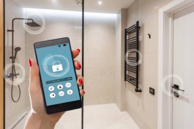 Elinde akıllı telefon tutan ve evinde güvenlik uygulaması olan genç bir kadın. Ev güvenliğini mobil bir cihazdan kontrol etme ve yönetme kavramı.
