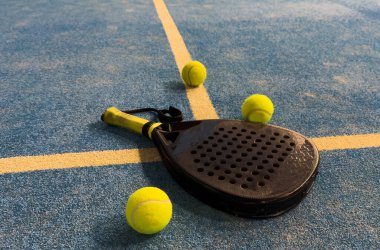 Bir padel veya raket tenis raketi ve parlak sarı bir tenis topu mavi bir kortta yatar. Ağ ve bir oyuncunun bulanık figürü arka planda görünür. Yüksek kalite fotoğraf
