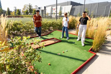 Bir kadın kızına golf oynamayı öğretiyor. Golf sahasında duruyorlar, kız bir golf sopası tutuyor, kadın arkasında duruyor ve ona vurmasını söylüyor. Yüksek kalite fotoğraf