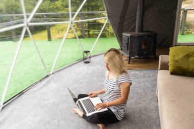 Glamping 'de şeffaf kabarcık çadırı, etrafındaki ve içindeki yemyeşil orman. Dizüstü bilgisayarlı kadın.