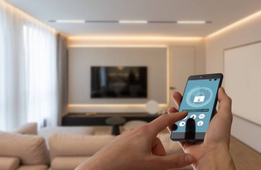 Elinde akıllı telefon tutan ve evinde güvenlik uygulaması olan genç bir kadın. Mobil bir cihazdan ev güvenliğini kontrol etme ve yönetme kavramı. Yüksek kalite fotoğraf