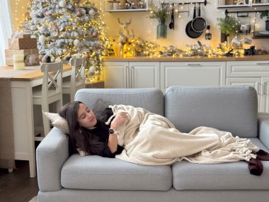 Neşeli kız, Noel ağacının yanındaki güzel oturma odasında bir kanepede yatıyor. Sıcak bir atmosferde battaniyeye sarılmış. Yüksek kalite fotoğraf
