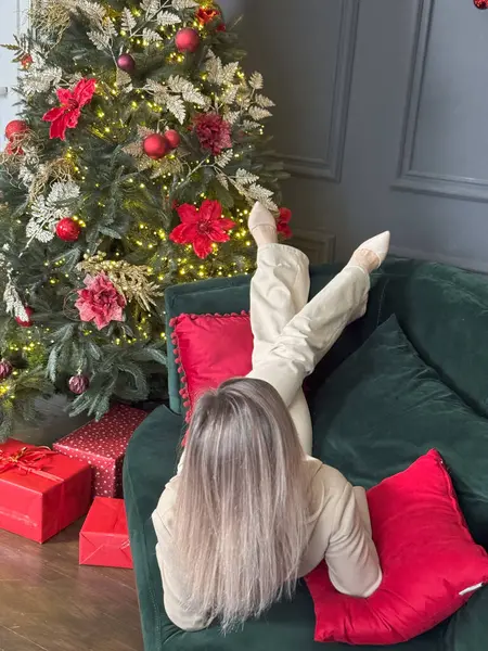 Sıcak bir tatil ortamında eğlenen bir kadın. Genç bir kadın, süslü bir Noel ağacıyla süslenmiş rahat bir oturma odasında dinleniyor. Yüksek kalite fotoğraf