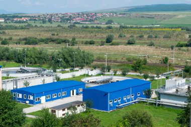 Kırsal alanda mavi yapıları ve tankları olan ve uzak bir köyü olan yeşil tepeleri yuvarlayan bir endüstri tesisi.