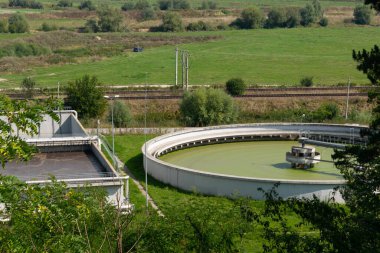 Daire ve dikdörtgen tankları olan bir su arıtma tesisinin hava görüntüsü kırsal bir alanda yeşil alanla çevrili.