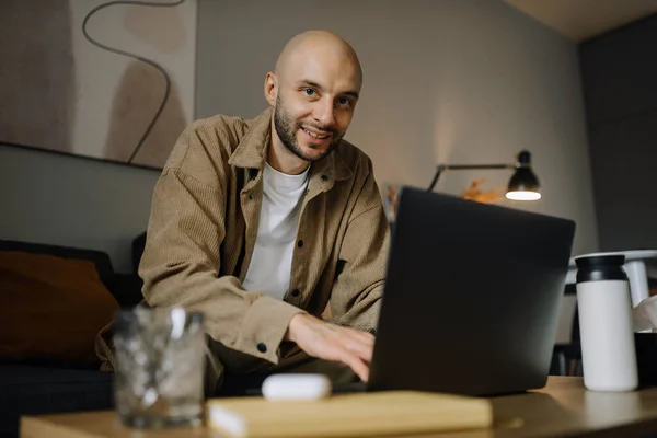 A smiling man is working on a laptop at home and smiled looking at us