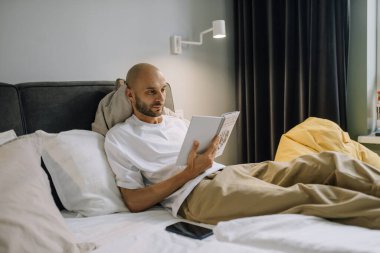 A man in home clothes is lying in a white bed in a bright bedroom reading a book in the afternoon