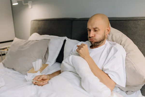 A man is sick and coughing in bed holding a thermometer