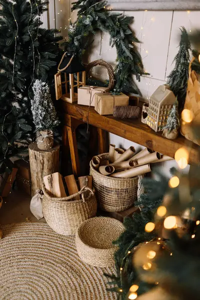 Masadaki Noel kompozisyonu: Çam çelenkleri, hasır sepet, dekoratif unsurlar ve bayram süslemeleri. Arka planda parıldayan çelenkler ve bir Noel ağacı görülebilir. İçerik için dikey fotoğraf