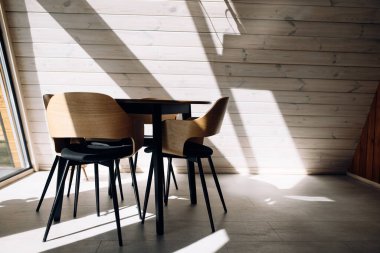 Parlak minimalist yemek alanı, ahşap masa ve güneş ışığında sandalyeler. İskandinav iç dizaynı açık tahta duvarlar ve gölgeler.
