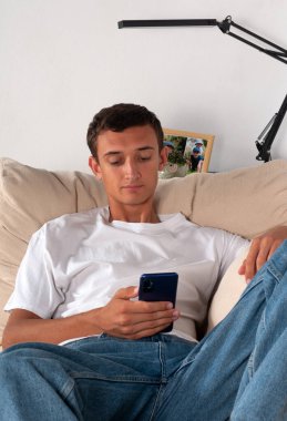 Cep telefonlu genç adam evdeki kanepede uzanıyor.