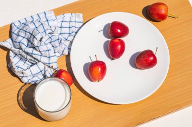 Yüksek açılı taze kırmızı, olgun, mini elma ya da beyaz seramik tabakta yengeç elması kahverengi ahşap kesme tahtasında bir bardak taze sütle doğrudan güneş ışığı altında..