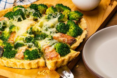 Yeni pişmiş lezzetli ev yapımı brokoli ve somonlu kiş ve tahta kesme tahtasının üzerinde beyaz seramik süt kabı olan dilimin yakın görüntüsü..