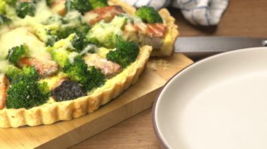 Taking a slice of freshly baked delicious homemade french food broccoli and salmon quiche from wooden cutting board and placing into brown ceramic dish on wooden table.