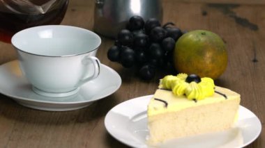 Pouring black coffee from transparent glass jar into white ceramic cup in saucer with delicious homemade sponge cake in white plate for breakfast with bunch of black grapes and an orange.
