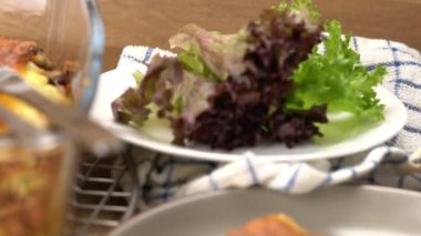 Moving over fresh vegetables in white ceramic plate, freshly baked delicious homemade italian food lasagna in transparent glass container on metal cooling rack and a piece of lasagna pouring with tomatoes sauce in brown ceramic dish.