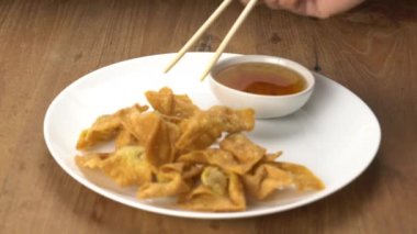 Using chopsticks clamp a crispy delicious homemade deep fried wonton from white ceramic dish, dip in sweet dipping sauce, and taking out.