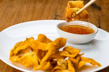 Using chopsticks clamp a delicious homemade crispy deep fried wonton in white ceramic dish dipping in sweet dipping sauce before eating.