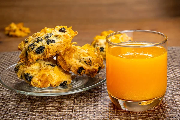 Şeffaf cam tabakta ev yapımı mısır gevreği kurabiyeleri ve siyah masa paspasında bir bardak portakal suyu..