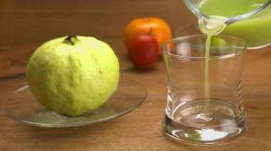 Şeffaf bardağa taze yeşil guava suyu dökülüyor cam tabakta olgun guava meyvesi portakal meyvesi ve ahşap masada olgun domates..