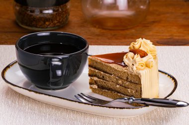 Bir fincan sade kahve ve krema ve bademle süslenmiş koyu kenarlı beyaz seramik tepsi ve ahşap masadaki metal çatalla süslenmiş bir dilim kahve..