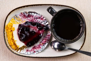 Folyo kağıdındaki yabanmersinli peynirli turta ve beyaz seramik tepside siyah seramik bardakta bir fincan kahve ve ahşap masadaki metal kaşık..