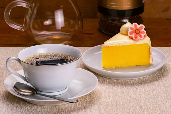 Beyaz seramik fincanda bir fincan sade kahvenin yanında beyaz seramik tabakta metal bir kaşık ve açık kahverengi masada beyaz seramik tabakta kremayla süslenmiş vanilyalı sünger keki..