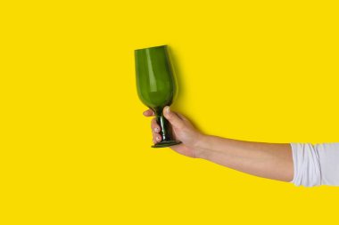 Sarı zemin üzerinde kocaman boş bir yeşil şarap kadehi tutan bir kadın.