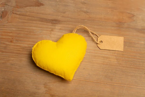 A fabric yellow heart with a cardboard label on a wooden background with copy space