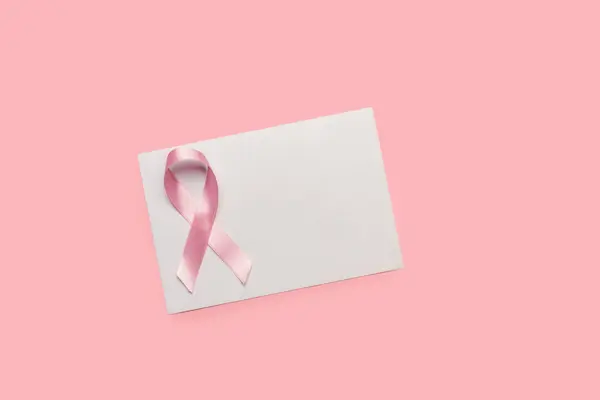 A pink ribbon bow on a white paper sheet on a pink background in a top view