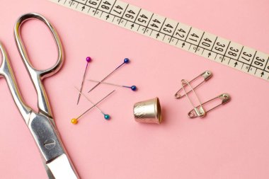 Makas, dikiş iğnesi, yüksük ve dikiş aksesuarları pembe arka planda.