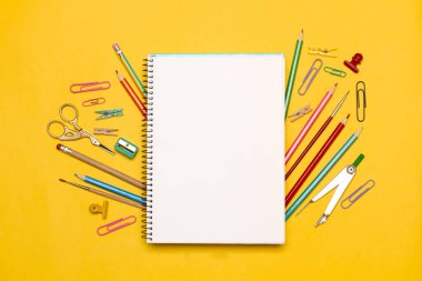 İçinde makas, renkli kalemler, ataçlar ve sarı arkaplanda bir pusula olan boş bir not defteri.