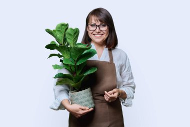 Beyaz stüdyo arkaplanındaki kameraya bakan önlüklü orta yaşlı kadın portresi. Bahçe düzenleme fabrikası. Çiçekçilik reklamı satıyor.