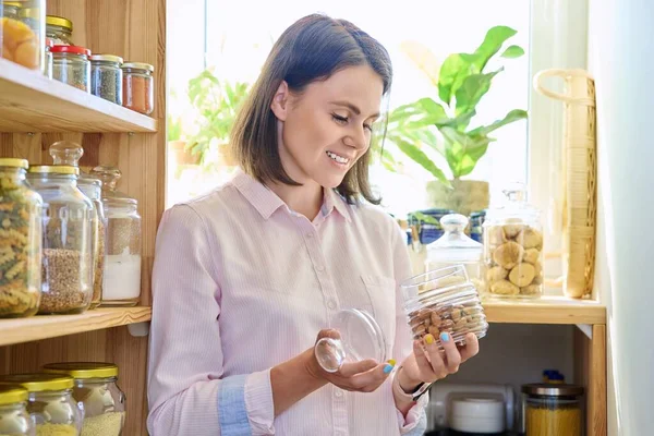 Young woman in kitchen with containers jars of cereals, pasta, dry fruits, holding jar of almonds. Female in home pantry, food organization and storage, kitchen utensils, household