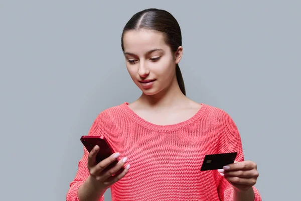Young teenage girl using smartphone and credit card, on grey color studio background. Adolescence, online shopping, food ordering, mobile applications, high school students, card payment concept