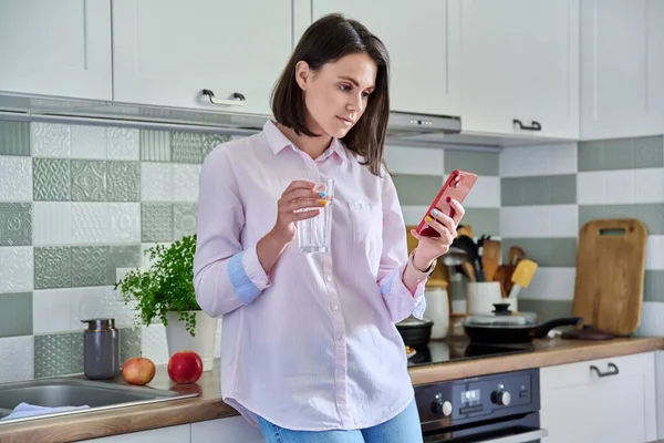 Young woman at home in kitchen looking in smartphone, with glass of water in hands. Beautiful smiling female drinking water, resting, using mobile apps, home interior background