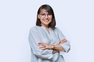 Beyaz stüdyo arka planında kameraya bakan kendinden emin, gülümseyen orta yaşlı bir kadın. Dost canlısı, olgun, saç stili makyajlı, gözlüklü, beyaz kolları çapraz beyaz gömlekli bir kadın..