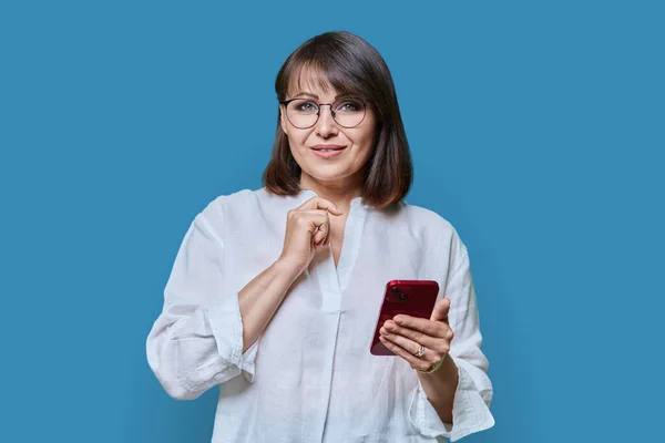 照片中快乐的中年美女 带着手机在蓝色的工作室背景上看着相机 穿着化妆型眼镜的成熟女性微笑 40人的科技生活方式 — 图库照片