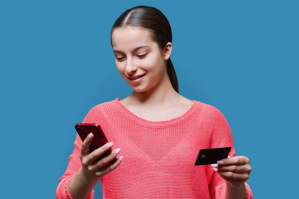 Young teenage girl using smartphone and credit card, on blue color studio background. Adolescence, online shopping, food ordering, mobile applications, high school students, card payment concept