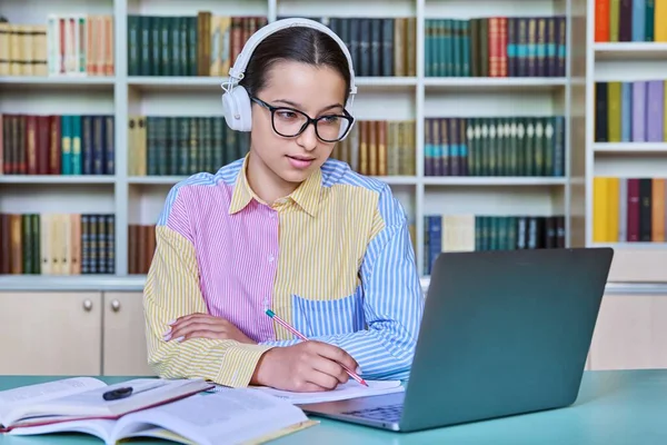 在图书馆学习的少女学生 女性在耳机中使用笔记本电脑 看着电脑 观看网络研讨会 在笔记本中做笔记 青少年 高中概念 — 图库照片
